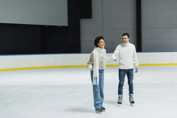 Positive interracial couple ice skating and holding hands during date on rink — Stock Photo