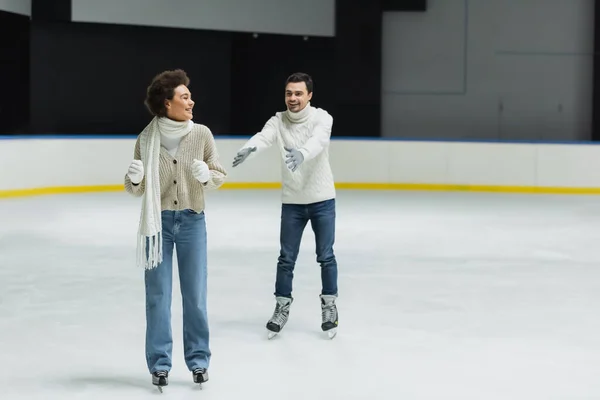 Smiling man outstretching hands to african american girlfriend ice skating on rink — Stock Photo