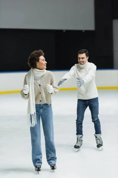 Sonriente mujer afroamericana patinaje sobre hielo cerca borrosa novio en pista de patinaje - foto de stock