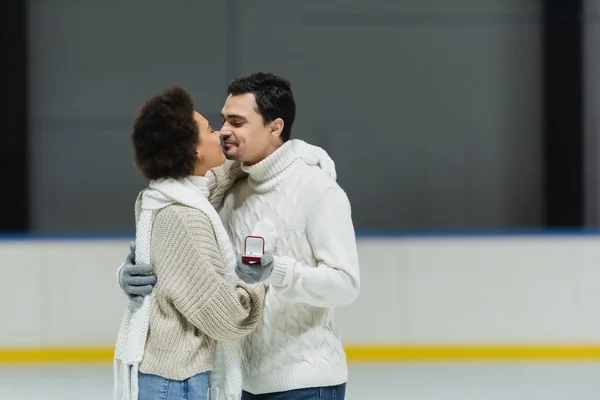 Jeune homme tenant la bague de demande en mariage et embrassant la petite amie afro-américaine sur la patinoire — Photo de stock