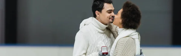 Man in sweater holding proposal ring and kissing african american girlfriend on ice rink, banner — Stock Photo