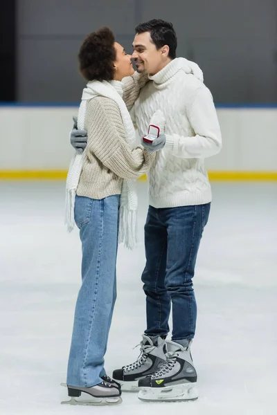 Vista lateral do casal multiétnico com proposta anel beijando na pista de gelo — Fotografia de Stock
