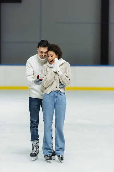Sonriente hombre sosteniendo anillo de compromiso cerca excitada afroamericana novia en pista de hielo - foto de stock