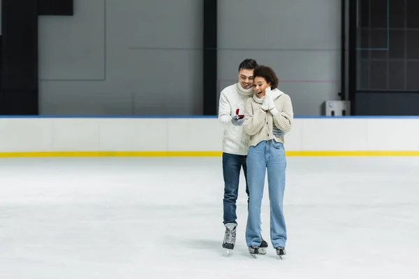Hombre sosteniendo anillo de propuesta y abrazando novia afroamericana en pista de hielo - foto de stock
