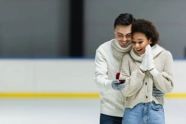 Junger Mann im Pullover hält Verlobungsring und umarmt afrikanisch-amerikanische Freundin auf Eisbahn — Stockfoto