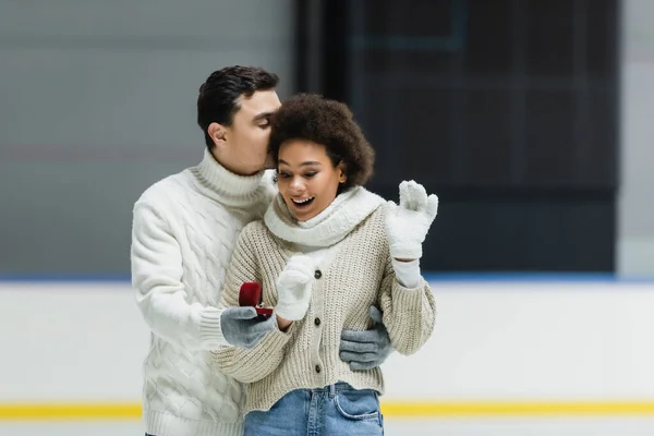 Hombre en suéter besar afro-americana novia y celebración propuesta anillo en pista de hielo - foto de stock
