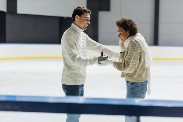 Vista laterale dell'uomo sorridente che mostra l'anello di fidanzamento alla ragazza afroamericana sulla pista di pattinaggio sul ghiaccio — Foto stock