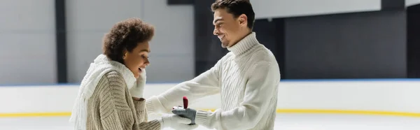 Side view of smiling man in sweater giving proposal ring to african american girlfriend on ice rink, banner — Stock Photo