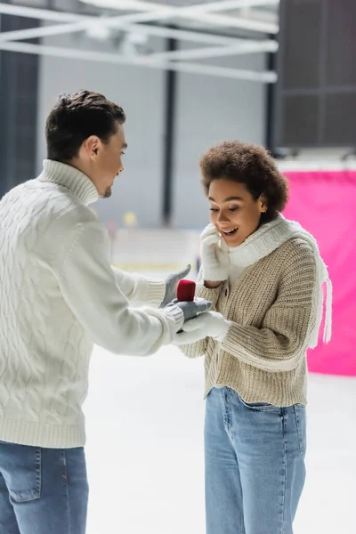 Young man holding proposal ring during date with african american girlfriend on ice rink — Stock Photo