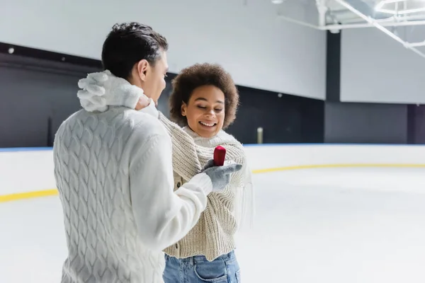 Cheerful african american woman embracing boyfriend with proposal ring on ice rink — Stock Photo
