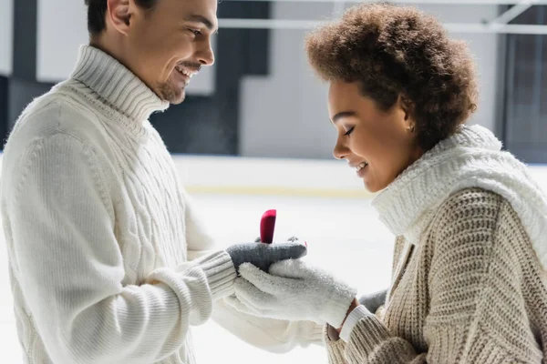 Side view of man in sweater holding engagement ring near african american girlfriend on ice rink — Stock Photo