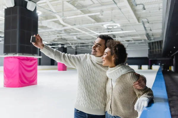 Smiling man taking selfie with african american girlfriend on ice rink — Stock Photo