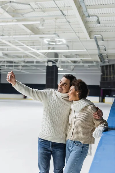 Smiling man hugging african american girlfriend and taking selfie on ice rink — Stock Photo