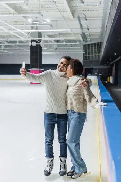 Smiling multiethnic couple taking selfie on smartphone on ice rink — Stock Photo