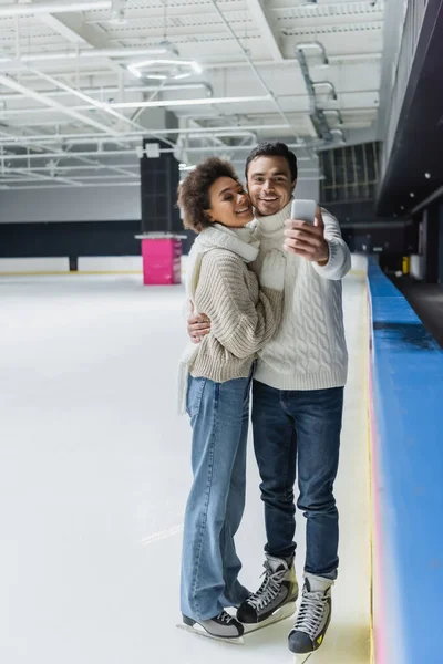 Jeune couple interracial en patins à glace et vêtements chauds prenant selfie sur la patinoire — Photo de stock