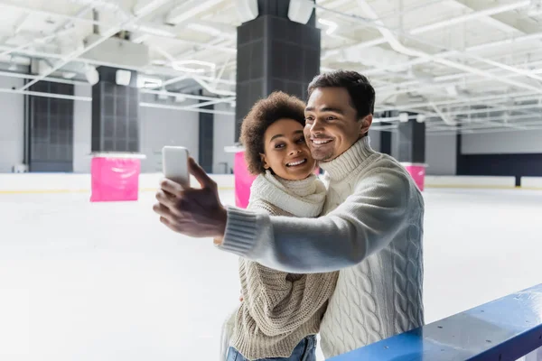 Positivo coppia multietnica prendendo selfie su smartphone sulla pista di pattinaggio di ghiaccio — Foto stock