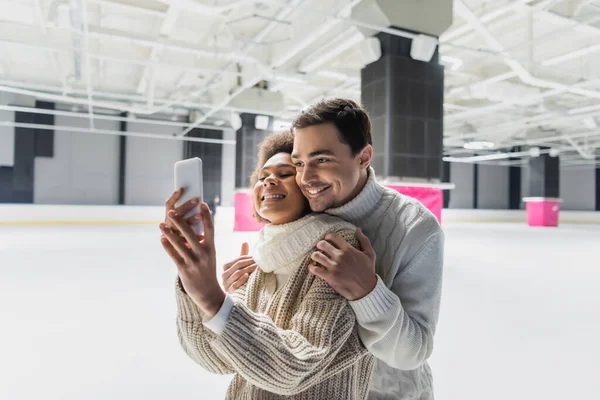 Hombre sonriente abrazando a novia afroamericana tomando selfie en smartphone en pista de hielo - foto de stock