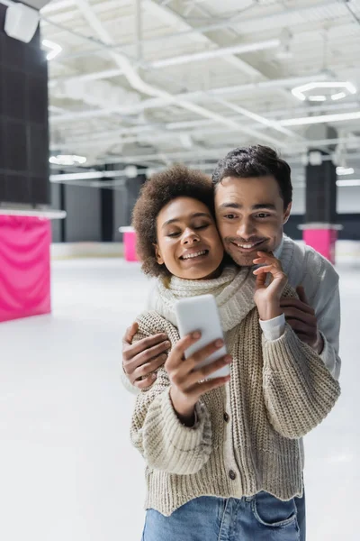 Smiling african american woman touching boyfriend and taking selfie on smartphone on ice rink — Stock Photo