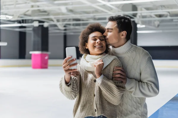 Joven besando a novia afroamericana tomando selfie en pista de hielo - foto de stock