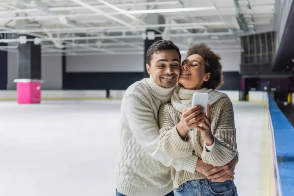 Uomo positivo in maglione che abbraccia la ragazza afroamericana con smartphone sulla pista di pattinaggio — Foto stock