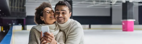 Mann umarmt afrikanisch-amerikanische Freundin mit Handy auf Eisbahn, Banner — Stockfoto