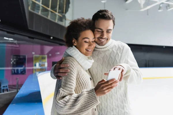 Lächelnder Mann umarmt afrikanisch-amerikanische Freundin und benutzt Smartphone auf Eisbahn — Stockfoto