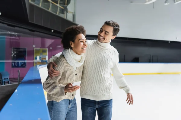 Positive man in sweater hugging african american girlfriend with smartphone on ice rink — Stock Photo