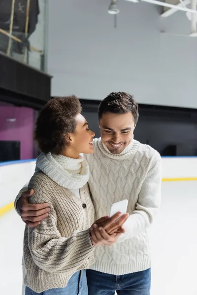 Smiling interracial couple hugging and using smartphone on ice rink — Stock Photo