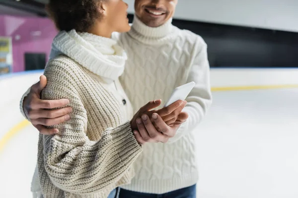 Cropped view of young multiethnic couple in warm clothes hugging and using smartphone on ice rink — Stock Photo