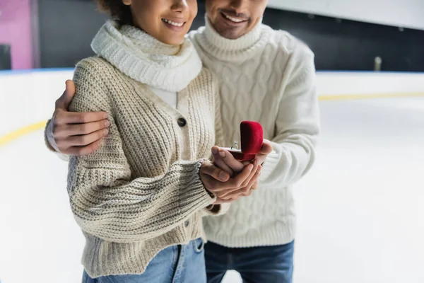 Vista recortada de hombre sonriente abrazando novia afroamericana con anillo de propuesta en pista de hielo - foto de stock