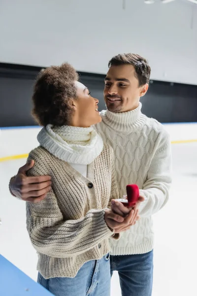 Smiling african american woman looking at boyfriend with blurred proposal ring on ice rink — Stock Photo
