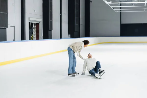 African american woman standing near cheerful boyfriend sitting on ice rink — Stock Photo