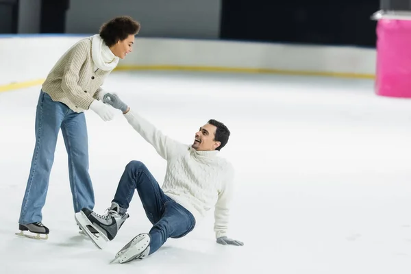 Sonriente mujer afroamericana ayudando novio cayendo en pista de hielo - foto de stock