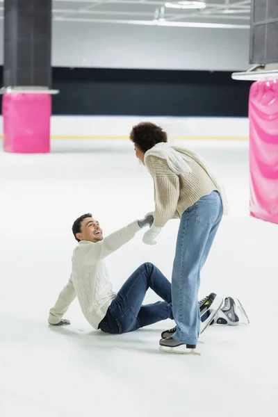 Afro americano donna in maglione aiutare fidanzato su ghiaccio pista di pattinaggio — Foto stock