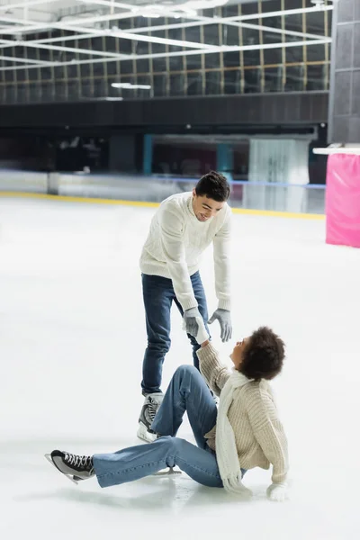 Young man giving hand to african american girlfriend falling on ice rink — Stock Photo