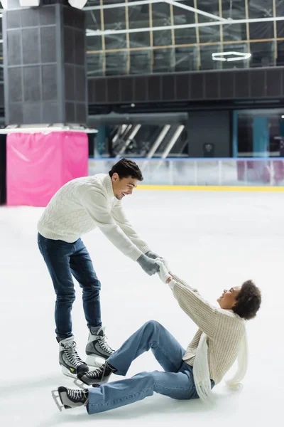 Side view of smiling man helping to african american girlfriend on ice rink — Stock Photo