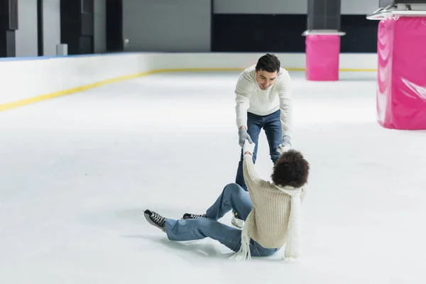 Young man in sweater helping to african american girlfriend on ice rink — Stock Photo