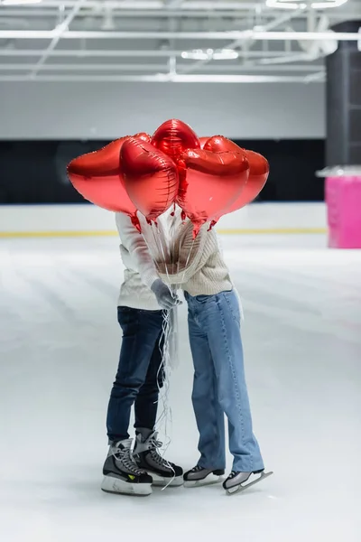 Jovem casal em roupas quentes segurando balões em forma de coração na pista de gelo — Fotografia de Stock