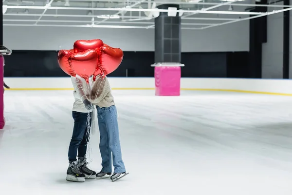 Pareja abrazando y sosteniendo globos rojos en forma de corazón en pista de hielo - foto de stock