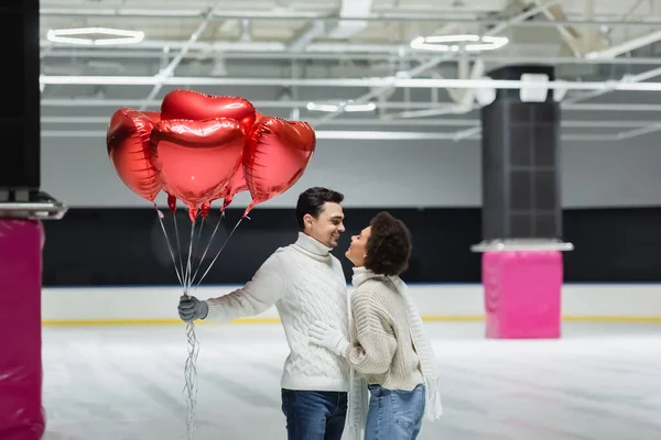 Vista lateral da mulher afro-americana sorridente abraçando namorado com balões em forma de coração na pista de gelo — Fotografia de Stock