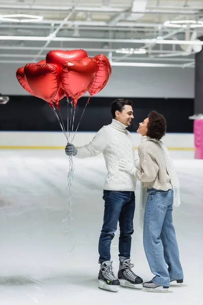 Vista lateral do homem sorridente segurando balões em forma de coração perto da namorada afro-americana na pista de gelo — Fotografia de Stock