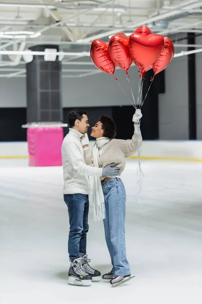 Vista lateral de la alegre mujer afroamericana sosteniendo globos en forma de corazón y tocando novio en pista de hielo - foto de stock