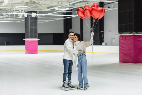 Pareja interracial positiva abrazando y sosteniendo globos en forma de corazón en pista de hielo - foto de stock