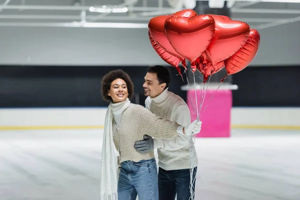 Lächelnder Mann im Pullover umarmt afrikanisch-amerikanische Freundin mit Luftballons in Herzform auf Eisbahn — Stockfoto