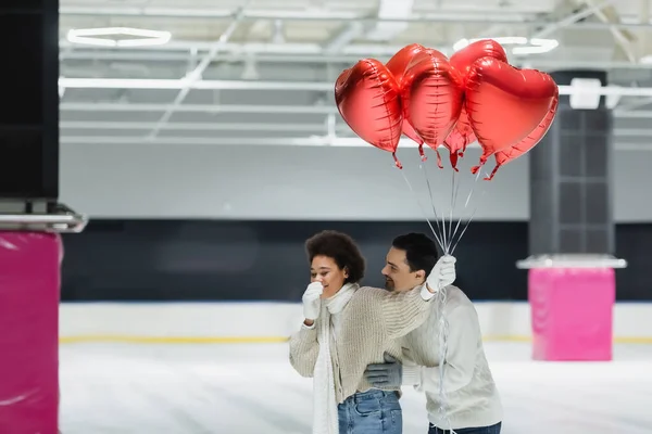 Young man hugging cheerful african american girlfriend with balloons in heart shape on ice rink — Stock Photo
