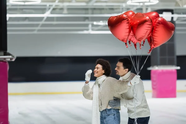 Giovane uomo che abbraccia allegra ragazza afro-americana in maglione con palloncini rossi a forma di cuore sulla pista di pattinaggio — Foto stock