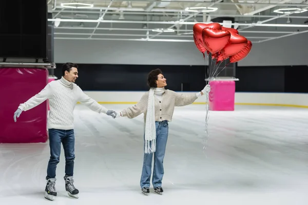 Visão lateral de casal interracial sorridente segurando balões em forma de coração enquanto patinação no gelo na pista — Fotografia de Stock