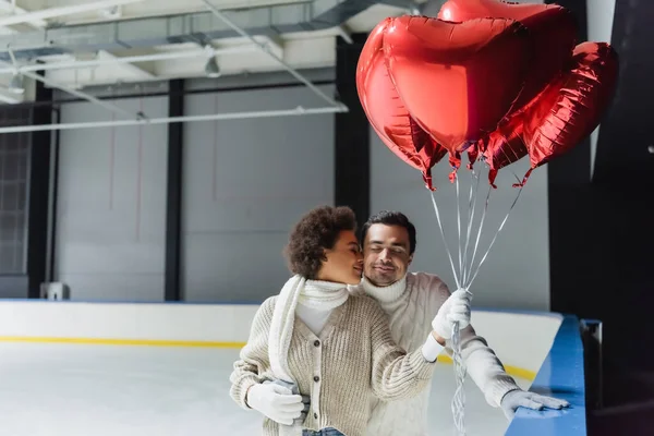 African american woman kissing boyfriend and holding heart shaped balloons on ice rink — Stock Photo
