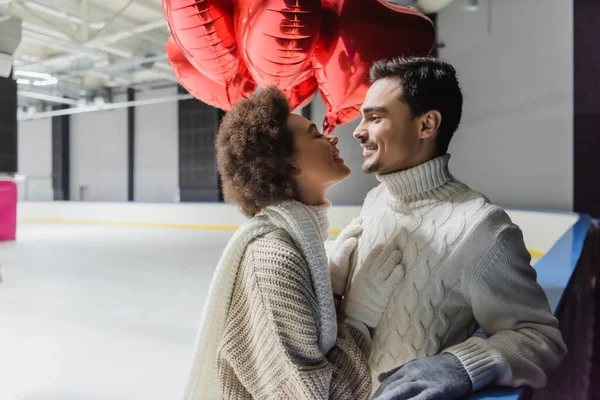 Vista lateral de la sonriente mujer afroamericana mirando al novio cerca de globos en forma de corazón en pista de hielo - foto de stock