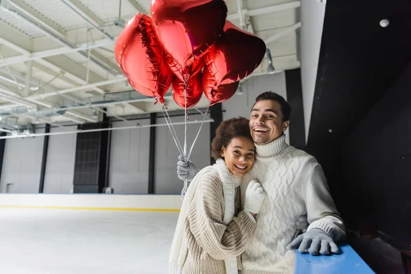 Alegre casal interracial segurando balões em forma de coração e olhando para a câmera na pista de gelo — Fotografia de Stock